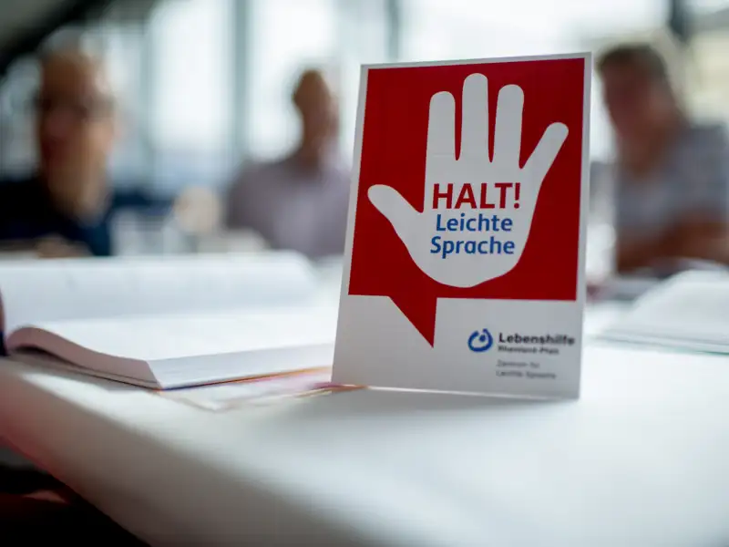 Auf einem Tisch steht ein Schild mit einer warnenden Hand und dem Hinweis: Halt! Leichte Sprache. Im Hintergrund sind unscharf drei Personen zu sehen.