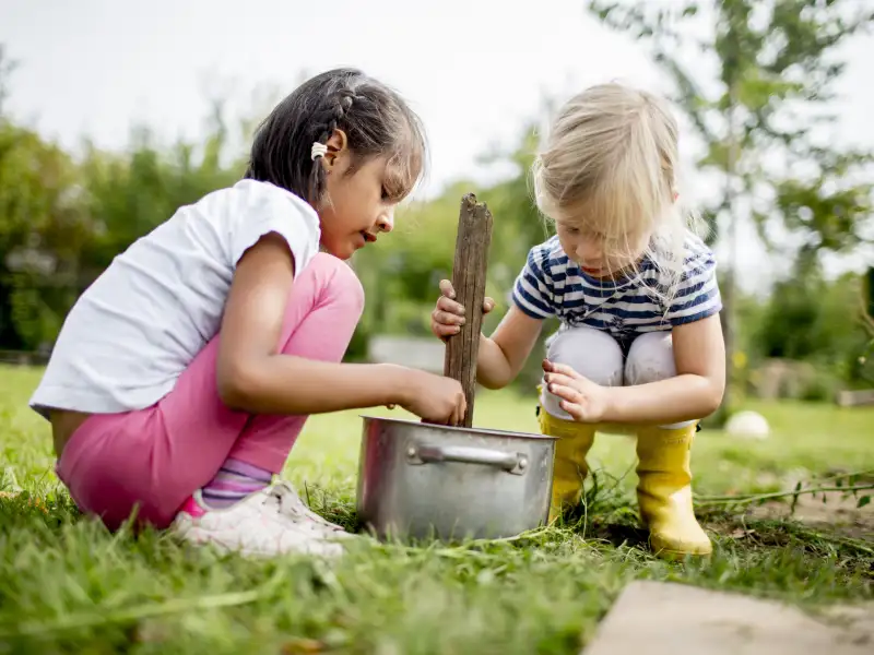 Zwei Kinder hocken im Gras vor einem Topf. Das eine Kind lässt etwas in den Topf fallen. Das andere Kind rührt mit einem Holzstock im Topf.