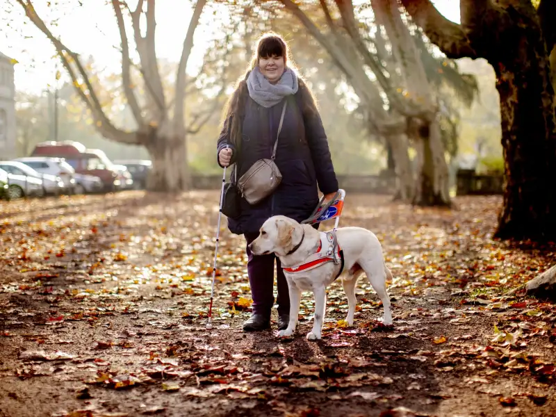 Ein Frau steht mit ihrem Blindenhund auf einem Fußweg.