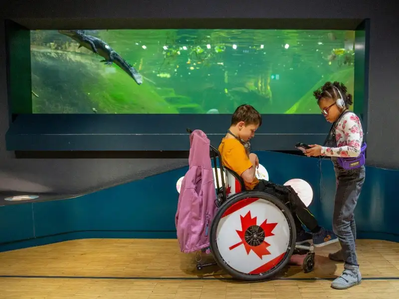 Ein Junge im Rollstuhl und ein Mädchen mit Kopfhörer stehen vor einem Aquarium.