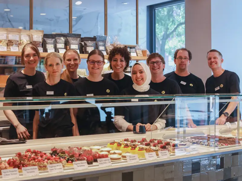 Eine Gruppe von Mitarbeitenden einer Konditorei steht hinter der Theke und lacht in die Kamera. Die Personen sind unterschiedlicher ethnischer Herkunft und mit und ohne sichtbare Behinderung.