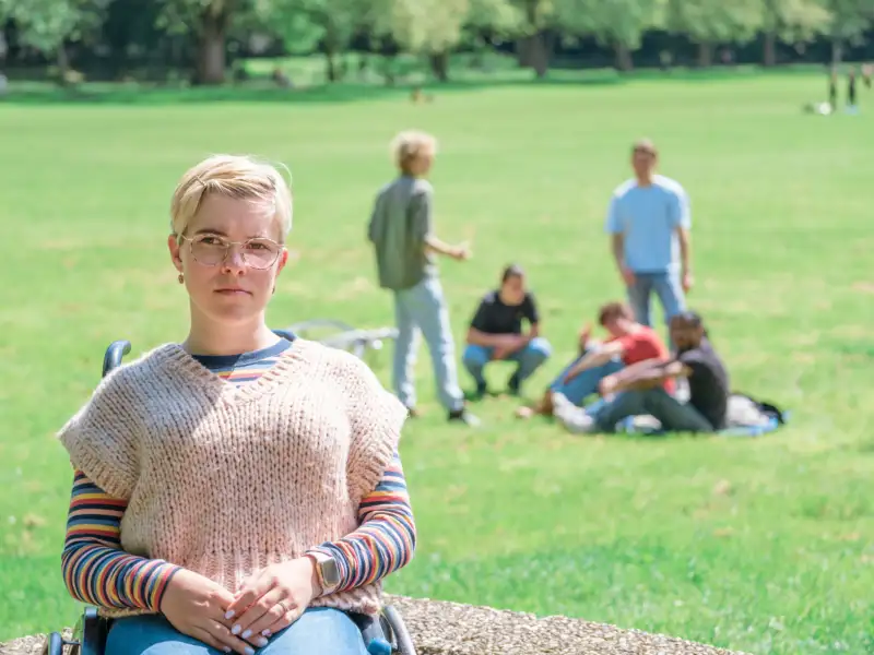 Eine junge Frau im Rollstuhl schaut in die Kamera. Im Hintergrund sieht man auf einer Wiese mehrere Jungedliche.