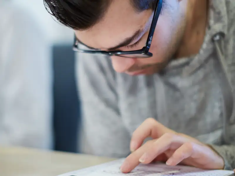 Ein junger Mann mit Brille beugt sich über ein Papier, das auf einem Tisch liegt. Seinen Finger nutzt er als Lesehilfe.
