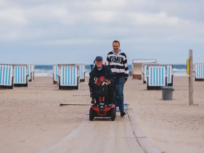 Zwei Männer gehen an einem Strand entlang. Einer der beiden nutzt einen Rollstuhl. Im Hintergrund sieht man das Meer sowie Strandkörbe.