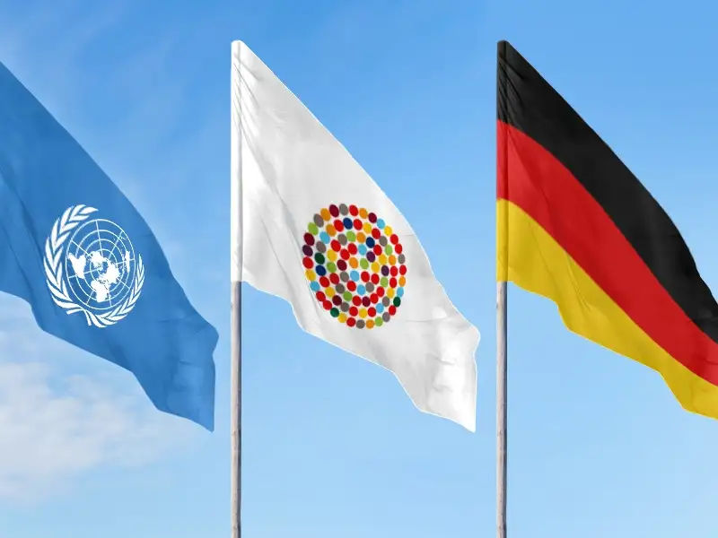 Drei Flaggen an Mast vor blauem Himmel: Eine mit dem Logo der UN, eine mit vielen bunten Punkten im Kreis, die Inklusion symbolisieren sollen und die deutsche Fahne