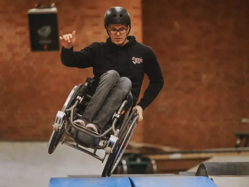 Ein Mann im Rollstuhl fährt mit einem Rollstuhlrad in einer Turnhalle über eine Matte. Das andere Rollstuhlrad berührt nicht den Boden.
