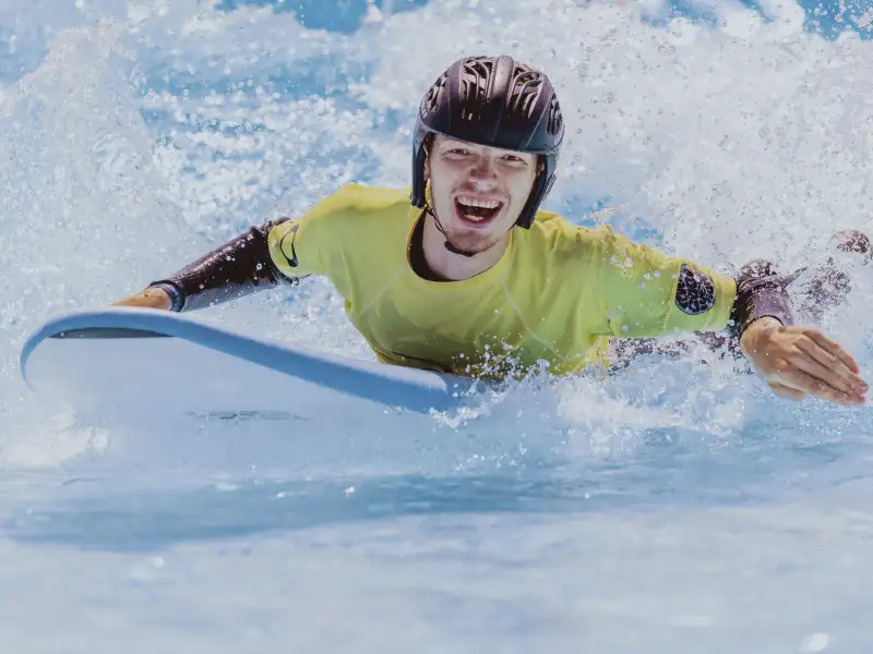 Ein junger Mann gleitet liegend auf einem Wellenbrett durch aufgewühltes Wasser. Er trägt einen Helm, Neoprenanzug und lacht begeistert in die Kamera.