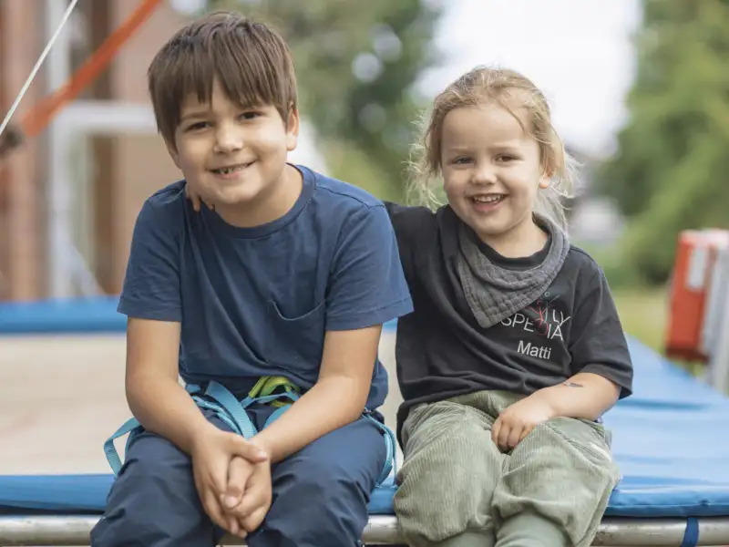 Zwei Jungs sitzen nebeneinander auf dem Rand eines Trampolins. Der kleinere hat seinen Arm freundschaftlich auf die Schulter des anderen gelegt. Beide tragen T-Shirts lächeln fröhlich in die Kamera.