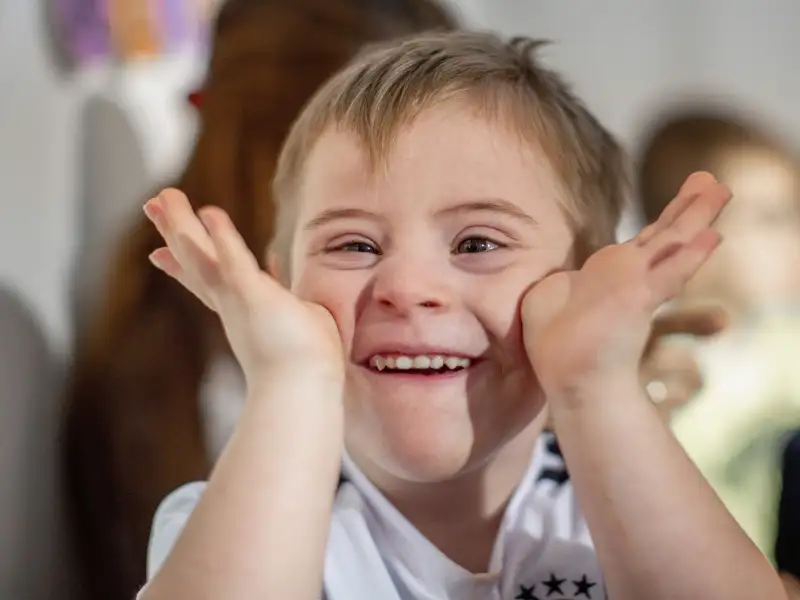 Ein kleiner Junge mit Down-Syndrom schneidet lachend eine Grimasse in die Kamera.