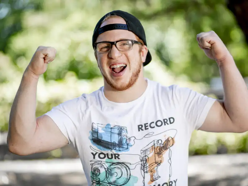 Ein junger Mann mit Behinderung zeigt lachend, wie stark er ist, indem er seine Arme nach oben anwinkelt und die Fäuste ballt. Er trägt eine Brille und ein verkehrtherum aufgesetztes Basecap.