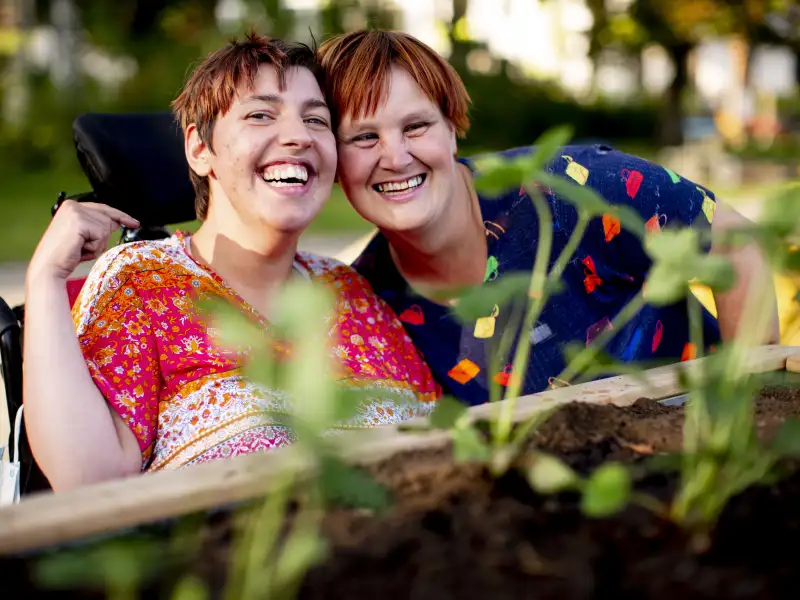 Zwei Frauen mit Behinderung sitzen hinter einem frisch bepflanzten Hochbeet. Eine der Frauen sitzt im Rollstuhl. Sie tragen sommerliche Kleidung und haben lachend die Köpfe aneinander gelegt. Beide schauen in die Kamera.