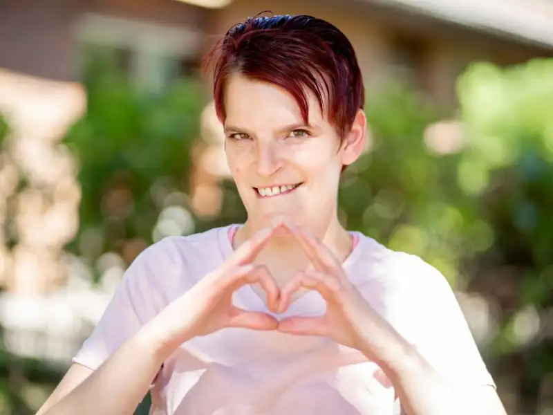 Eine junge Frau mit Behinderung zeigt lächelnd mit beiden Händen ein Herz-Symbol in die Kamera. Sie trägt kurzes rotes Harr und ein rosafarbenes T-Shirt.