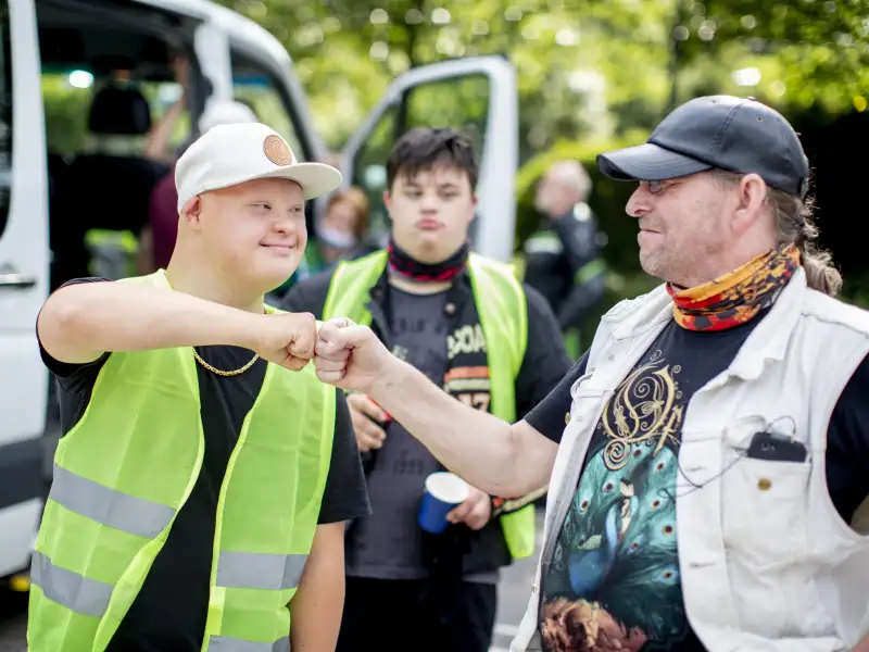 Zwei Biker begrüßen sich mit einem freundschaftlichen Faustgruß. Einer von ihnen hat das Down-Syndrom und trägt eine Warnweste. Der andere trägt ein schwarzes Basecap und ein buntes Halstuch.