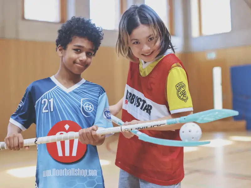 Zwei Jungen unterschiedlicher Hautfarbe balancieren gemeinsam einen Ball zwischen zwei Hockeyschlägern. Sie lachen in die Kamera.