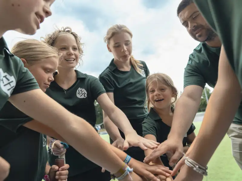 Eine Gruppe von Mädchen in grünen Sporttrikots mit ihrem Trainer. Sie schwören sich als Team ein, indem sie eine Runde bilden und alle ihre Hände aufeinandergelegt haben. Sie lächeln.