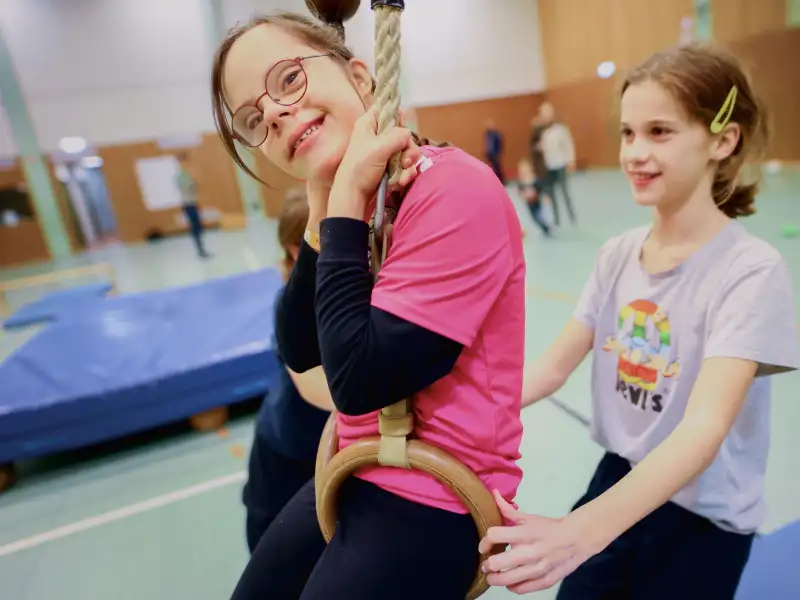 Zwei Mädchen in einer Turnhalle. Eine sitzt in Ringen, die andere schubst sie an.