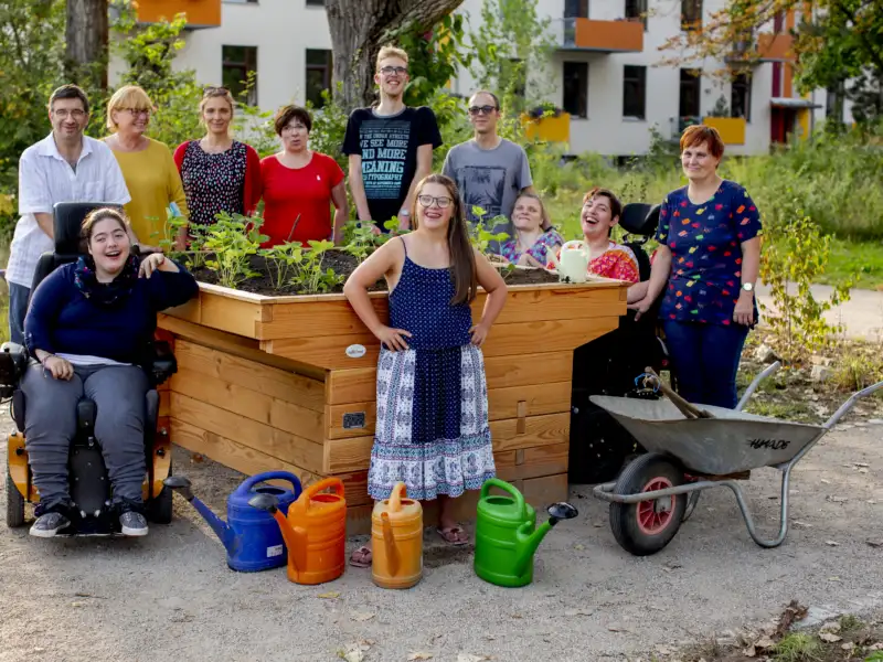 Eine Gruppe von Menschen mit und ohne Behinderung steht um ein Hochbeet, das sie gerade bepflanzt haben. Alle lächeln.