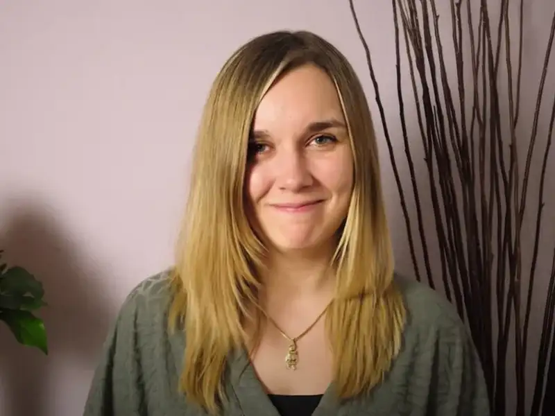 Eine junge Frau mit langen, blonden Haaren lächelt in die Kamera. Neben ihr steht ein Blumenstrauß.