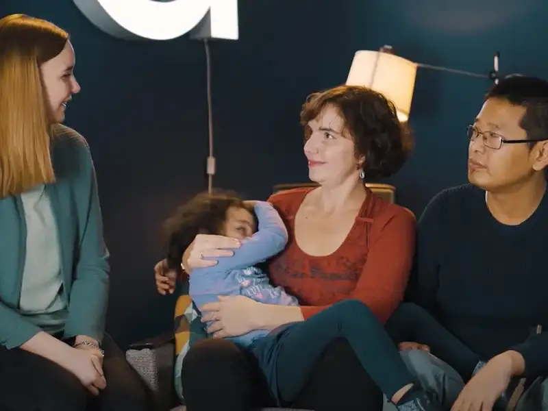 Drei blinde Menschen sitzen nebeneinander und schauen sich an. In der Mitte sitzt eine Frau, die ein vierjähriges Kind auf dem Schoß hat.