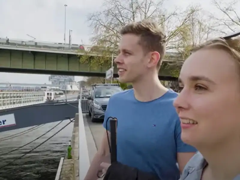 Ein junger Mann und eine junge Frau stehen am Rheinufer. Im Hintergrund sieht man eine Straßenbahn über eine Brücke fahren.