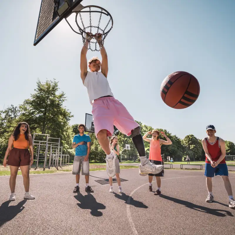Jugendliche auf einem Basketballplatz. Einer von ihnen trägt eine Beinprothese und hält sich am Basketballkorb fest.