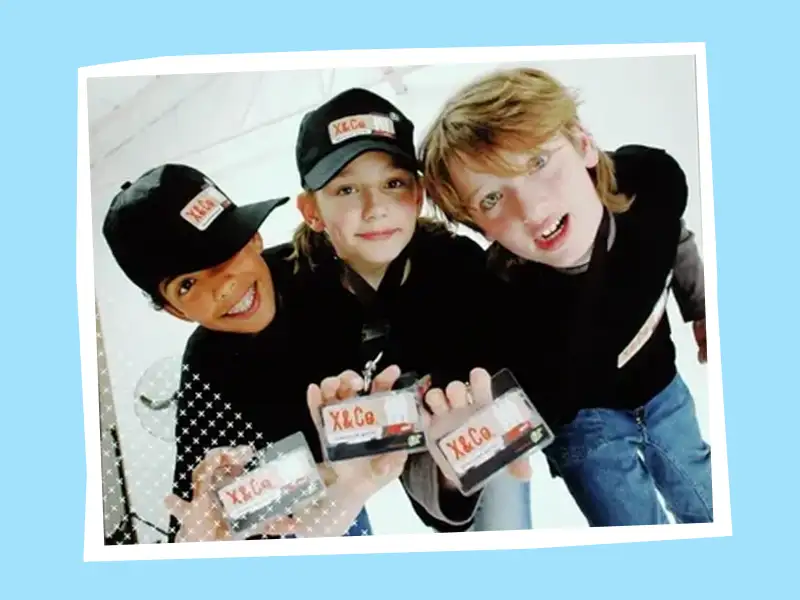 Drei Kinder mit schwarzen Cappies blicken in die Kamera u nd halten einen Ausweis mit dem Schriftzung XundCo. hoch.