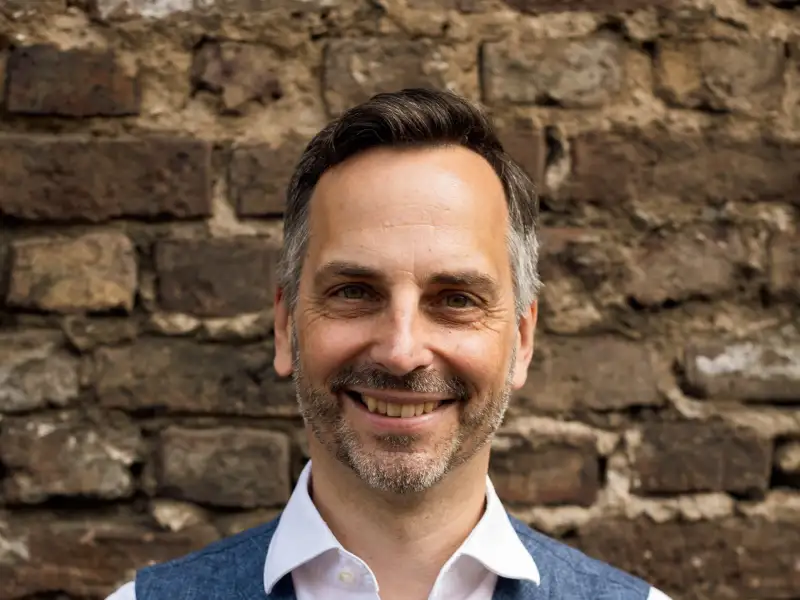 Portraitfoto von Frank Liffers. Er trägt kurze Haare und einen Dreitagebart und steht vor einer Backsteinmauer.