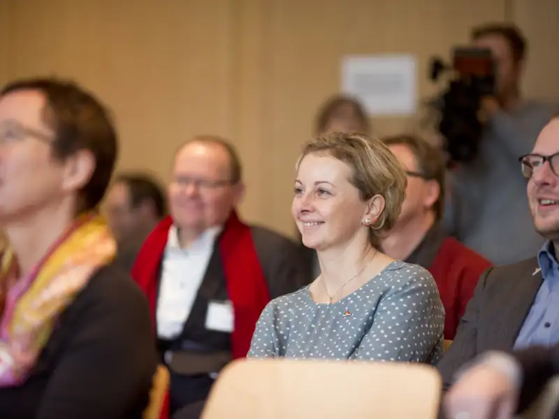 Teilnehmer*innen einer Konferenz sitzen in Stuhlreihen und schauen interessiert nach vorne.