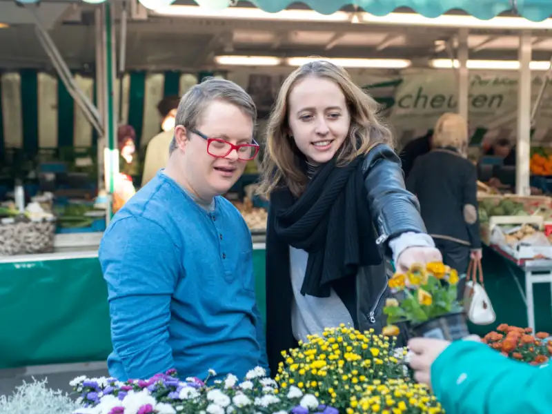 Ein junger Mann mit Down-Syndrom und eine junge Frau betrachten Blumen vor einem Marktstand.