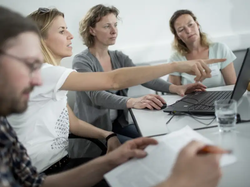 Mehrere Personen bei der gemeinsamen Arbeit an einem Laptop. Eine Frau zeigt mit dem Finger auf den Bildschirm.