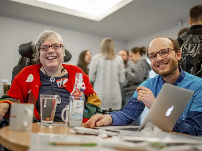 Eine Frau im Rollstuhl und ein Mann lachen in die Kamera.
