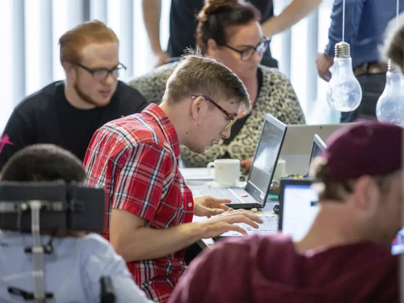 Ein junger Mann arbeitet an einem Laptop. Um ihn herum sitzen weitere Personen mit und ohne Behinderung und arbeiten ebenfalls.