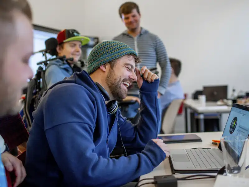 Ein Mann sitzt an einem Laptop. Er trägt einen Hoodie und eine Strickmütze und lacht. Im Hintergrund sind weitere Personen zu sehen, darunter ein junger Mann im Elektrorollstuhl.