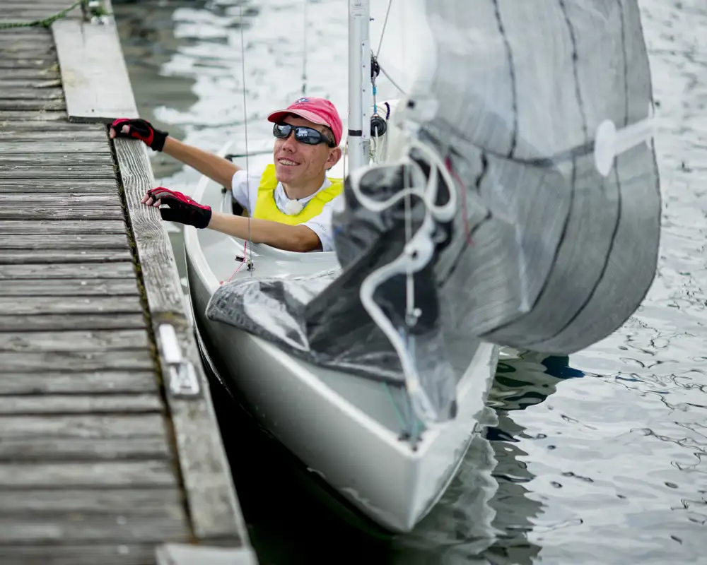 Basti, ein junger Mann mit Sonnenbrille und Basecap, sitzt in einem Segelboot und hält sich am Bootssteg fest.