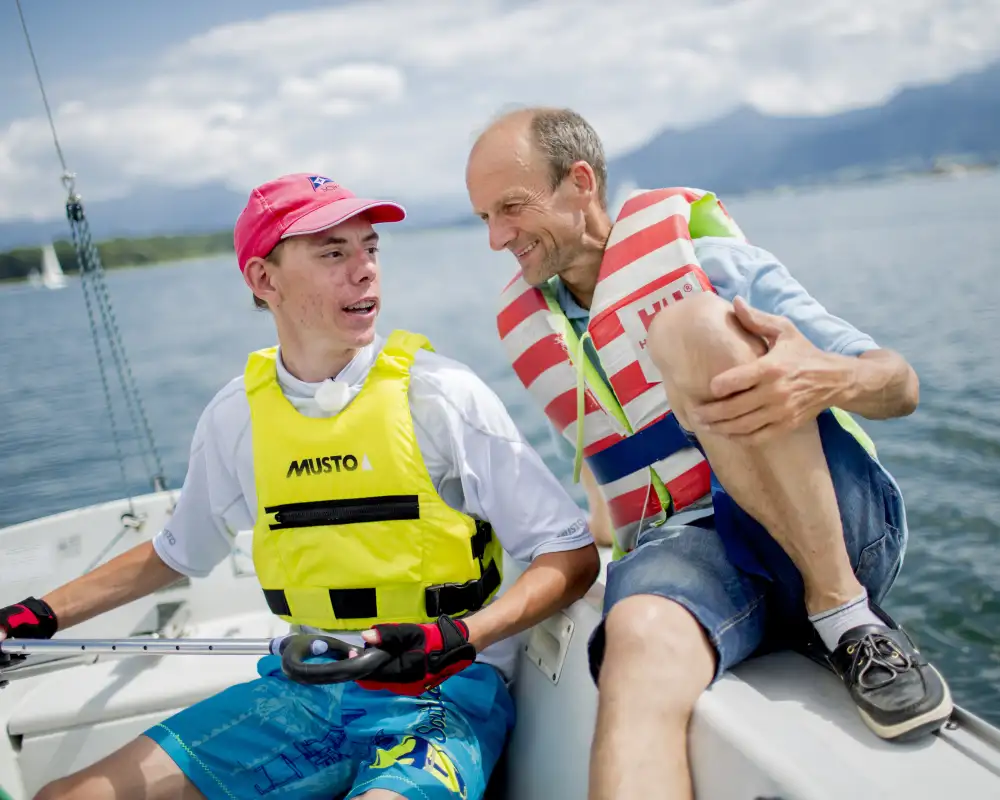 Basti, ein junger Mann mit Basecap und ein Mann sitzen nebeneinander auf einem Segelboot. Beide tragen Schwimmwesten. Im Hintergrund sieht man einen See.