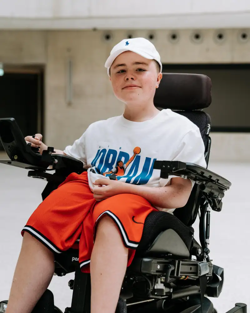 Ein Junge sitzt in einem elektrischen Rollstuhl. Er trägt Basketball-Klamotten und lächelt in die Kamera.