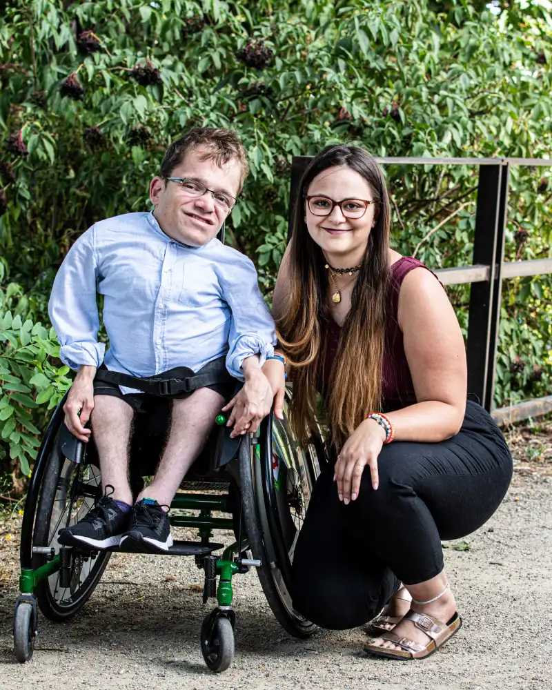 Cristina, eine junge Frau mit langen, dunklen Haaren und Brille hockt neben ihrem Partner, der kurze braune Haare hat, eine Brille trägt und im Rollstuhl sitzt.