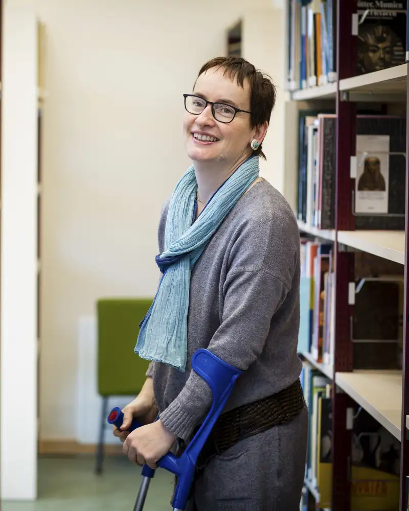 Agnes, eine 46-Jährige mit kurzen dunklen Haaren und Brille steht zwischen zwei Regalen.