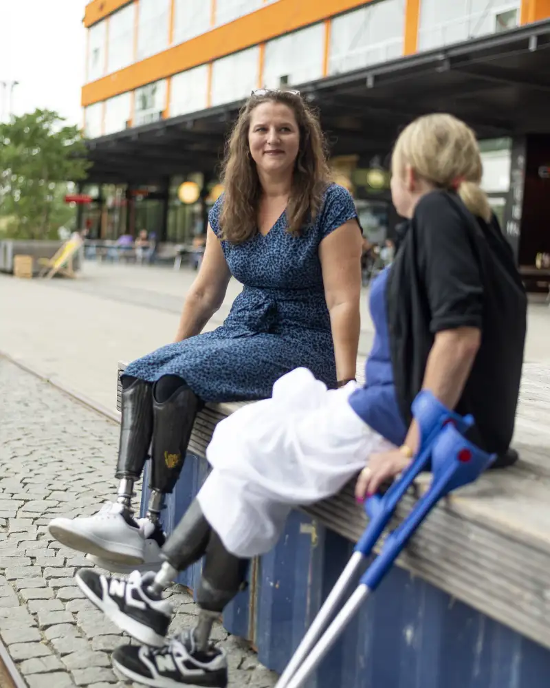 Zwei Frauen sitzen auf einem Mäuerchen und schauen sich an. Eine Frau sieht man von vorne, die andere von hinten. Beide tragen an beiden Beinen Prothesen.
