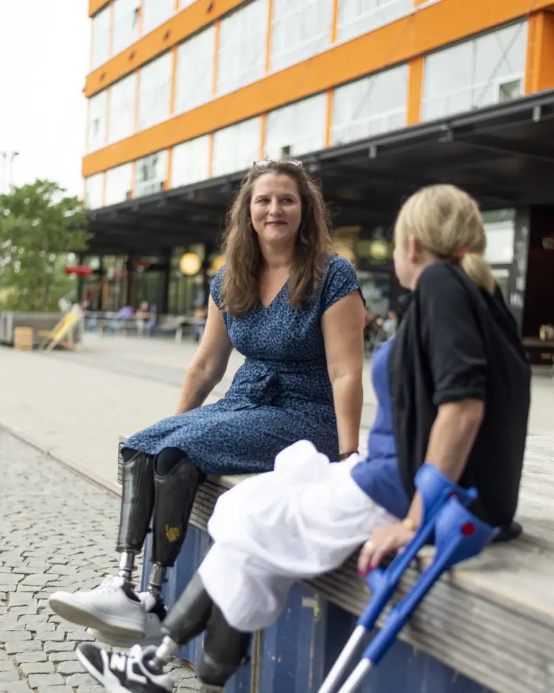 Zwei Frauen sitzen auf einem Mäuerchen und schauen sich an. Eine Frau sieht man von vorne, die andere von hinten. Beide tragen an beiden Beinen Prothesen.