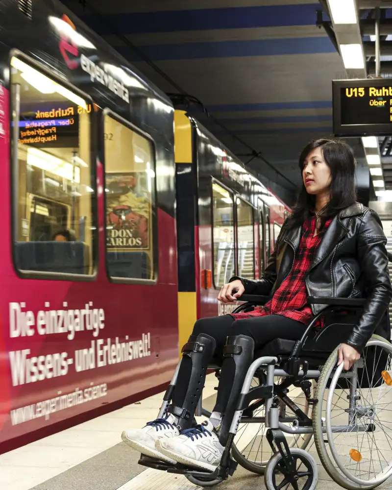 Simone, eine junge Frau mit langen dunklen Haaren im Rollstuhl, steht an einer U-Bahn-Haltestelle. Die Bahn ist gerade eingefahren.