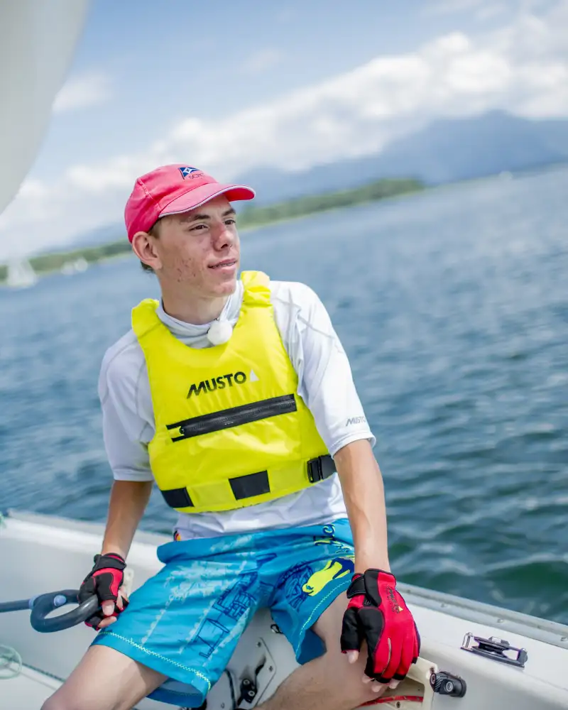 Basti, ein junger Mann mit kurzen Haaren sitzt in einem Segelboot. Er trägt ein rotes Basecap, ein weißes Shirt, blaue Shorts und eine Schwimmweste. Im Hintergrund sieht man einen See.