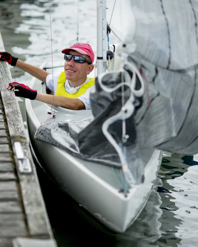 Basti, ein junger Mann mit Sonnenbrille und Basecap, sitzt in einem Segelboot und hält sich am Bootssteg fest.