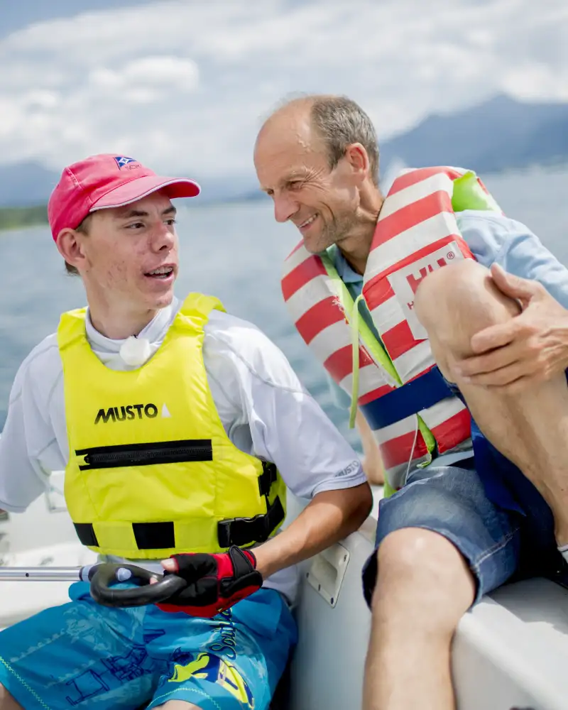 Basti, ein junger Mann mit Basecap und ein Mann sitzen nebeneinander auf einem Segelboot. Beide tragen Schwimmwesten. Im Hintergrund sieht man einen See.