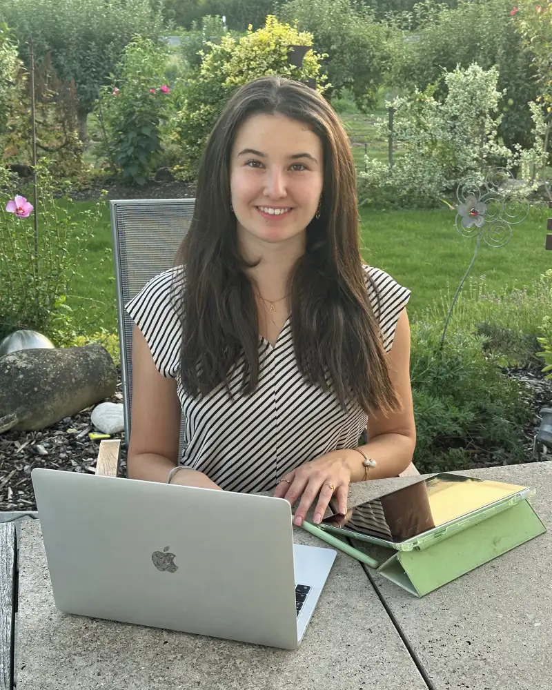Eine junge Frau mit langen braunen Haaren sitzt in einem Garten. Vor ihr auf dem Tisch stehen ein Laptop und ein Tablet.