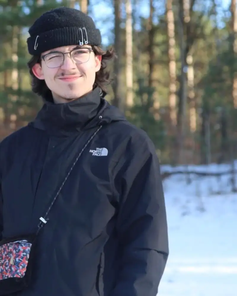Porträt eines jungen Mannes in einer Schneelandschaft. Er trägt eine Brille und eine Mütze. Ein Auge kneift er zu.