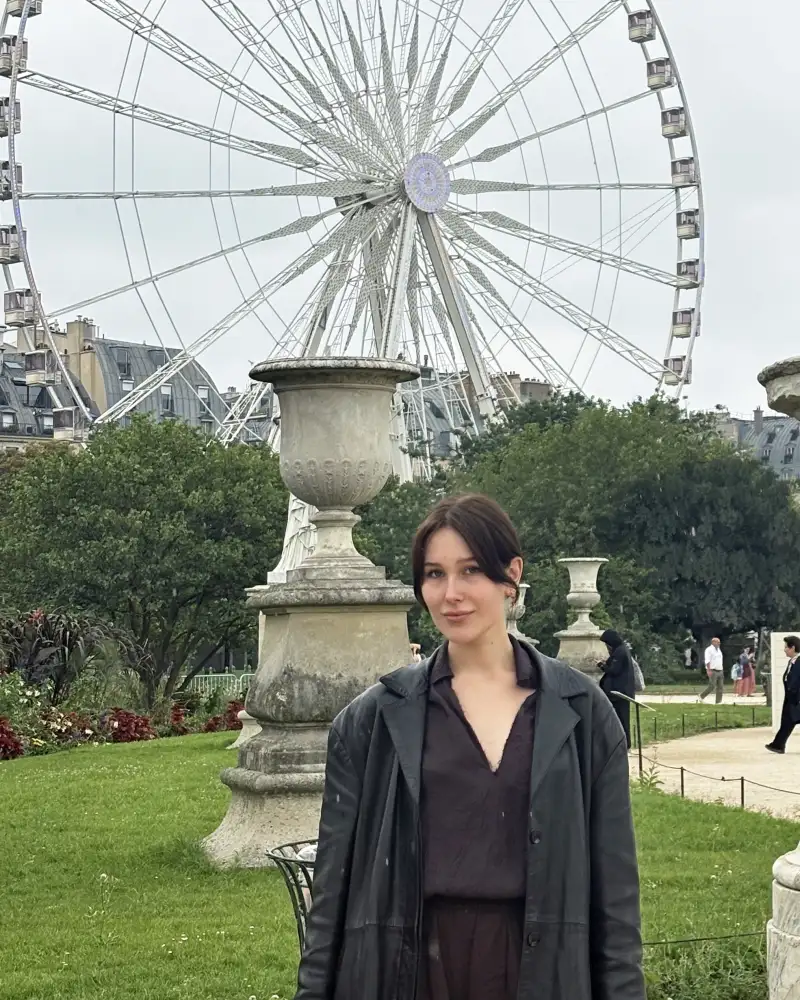 Eine junge Frau mit dunklen Haaren, die nach hinten frisiert sind steht in einem Park. Im Hintergrund sieht man ein Riesenrad.