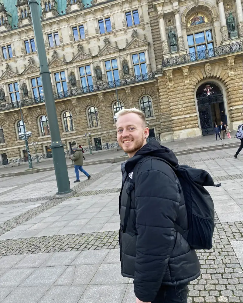 Ein junger Mann mit kurzen, blonden Haaren und kurzem Bart steht auf einem städtischen Platz vor einem historischen Gebäude.