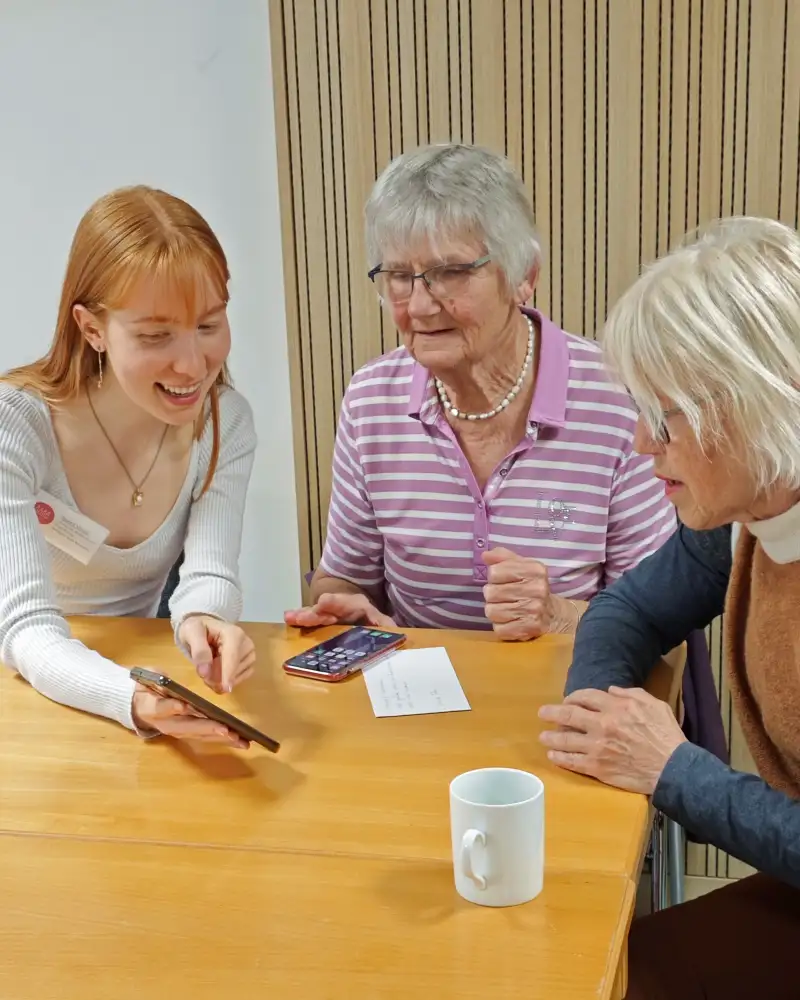 Eine junge Frau mit langem rötlichen Haar zeigt zwei Seniorinnen etwas auf einem Smartphone.