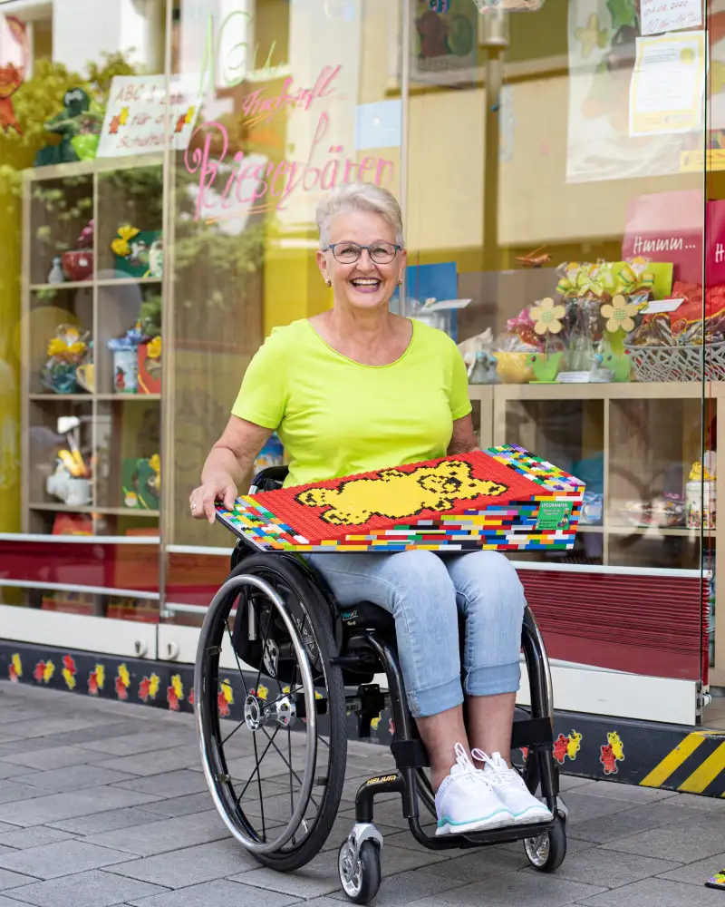 Eine Frau mit kurzen grauen Haaren und sommerlicher Kleidung sitzt in ihrem Rollstuhl vor einem Süßwarenladen und hält eine ihrer selbstgebauten Legorampen auf dem Schoß. Auf der Rampe sind bunte Gummibärchen abgebildet.